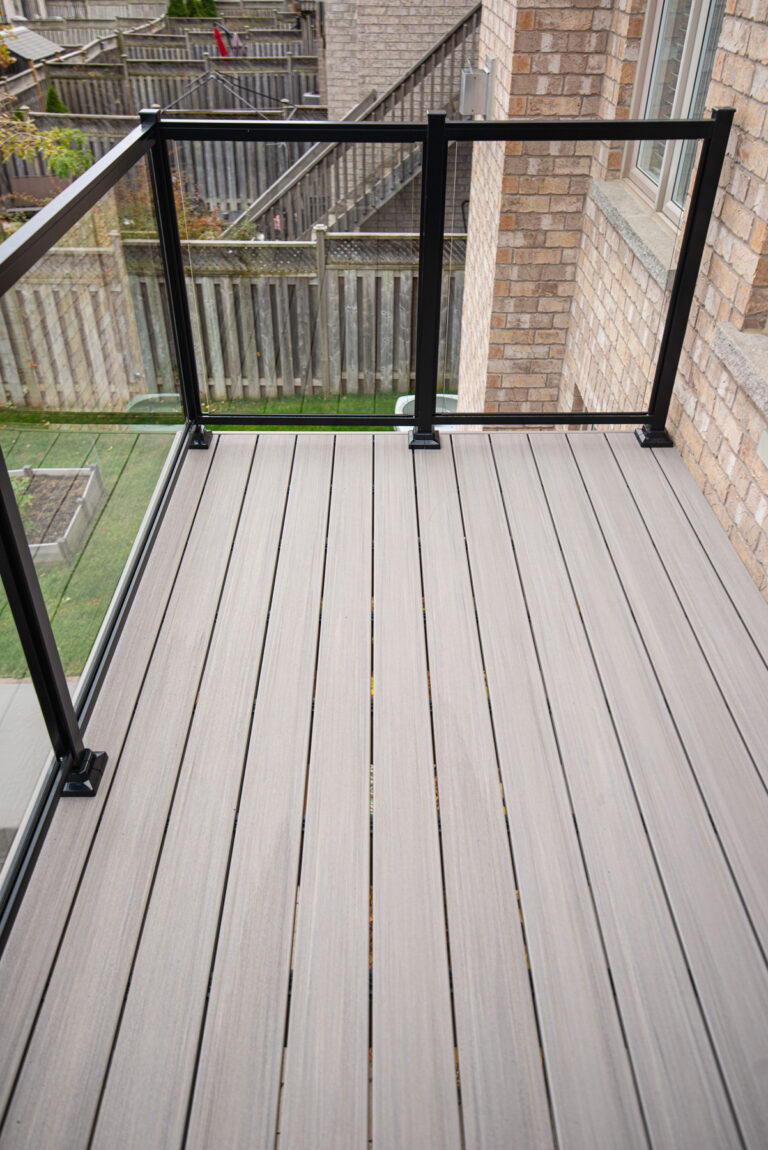 A modern balcony with light gray composite decking and glass-panel railings overlooking a backyard with a wooden fence and garden beds. Brick walls and windows are visible beside the balcony.