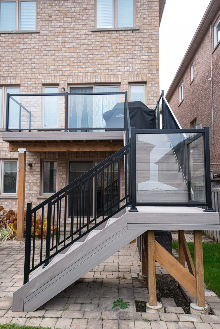 A modern backyard deck with black metal and glass railings, wooden support beams, and stairs leading down to a stone patio, attached to a brick house.