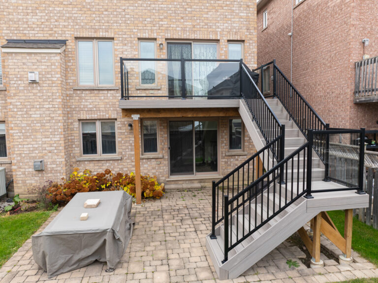 Backyard view of a brick house with a raised composite deck, black metal railings, and stairs leading to a paved patio. A covered outdoor table sits on the patio, with plants lining the house wall.