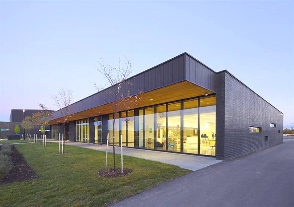 A modern, single-story building with large glass windows reflecting the evening sky, featuring a sleek black exterior and wood-toned ceiling. The surrounding area includes a paved path, grassy expanse, young leafless trees, and a spacious commercial deck for gatherings.