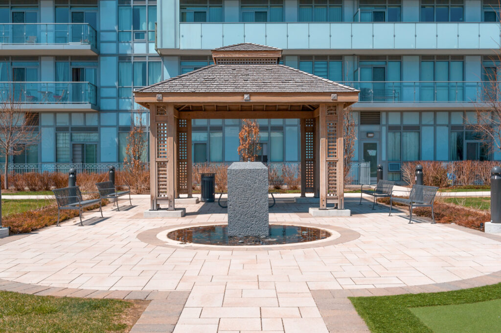 A wooden gazebo with a shingled roof, crafted by Delta Decks Toronto, stands on a paved area in front of a modern glass building. A stone fountain is at the center, surrounded by benches. Green grass and bare shrubs line the walkway.