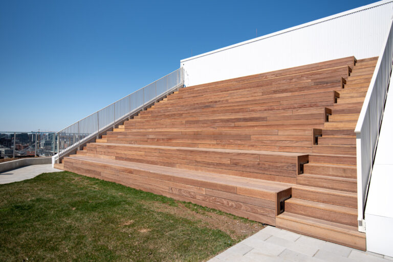 Outdoor wooden bleacher-style seating next to a grassy area, with a metal railing and a clear blue sky.