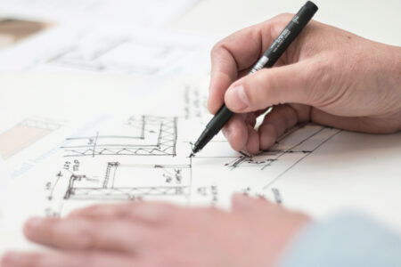 A persons hand holding a black pen sketches architectural drawings of a custom deck design on paper. The page tingles with detailed lines, measurements, and notes. The other hand rests on the Toronto plans, emphasizing precision and creativity in every stroke.