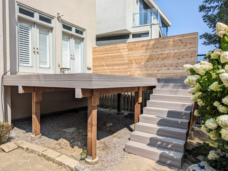 A raised wooden deck with stairs, supported by sturdy posts, is attached to a modern house. Theres a tall wooden privacy screen on one side and blooming white flowers in the foreground.