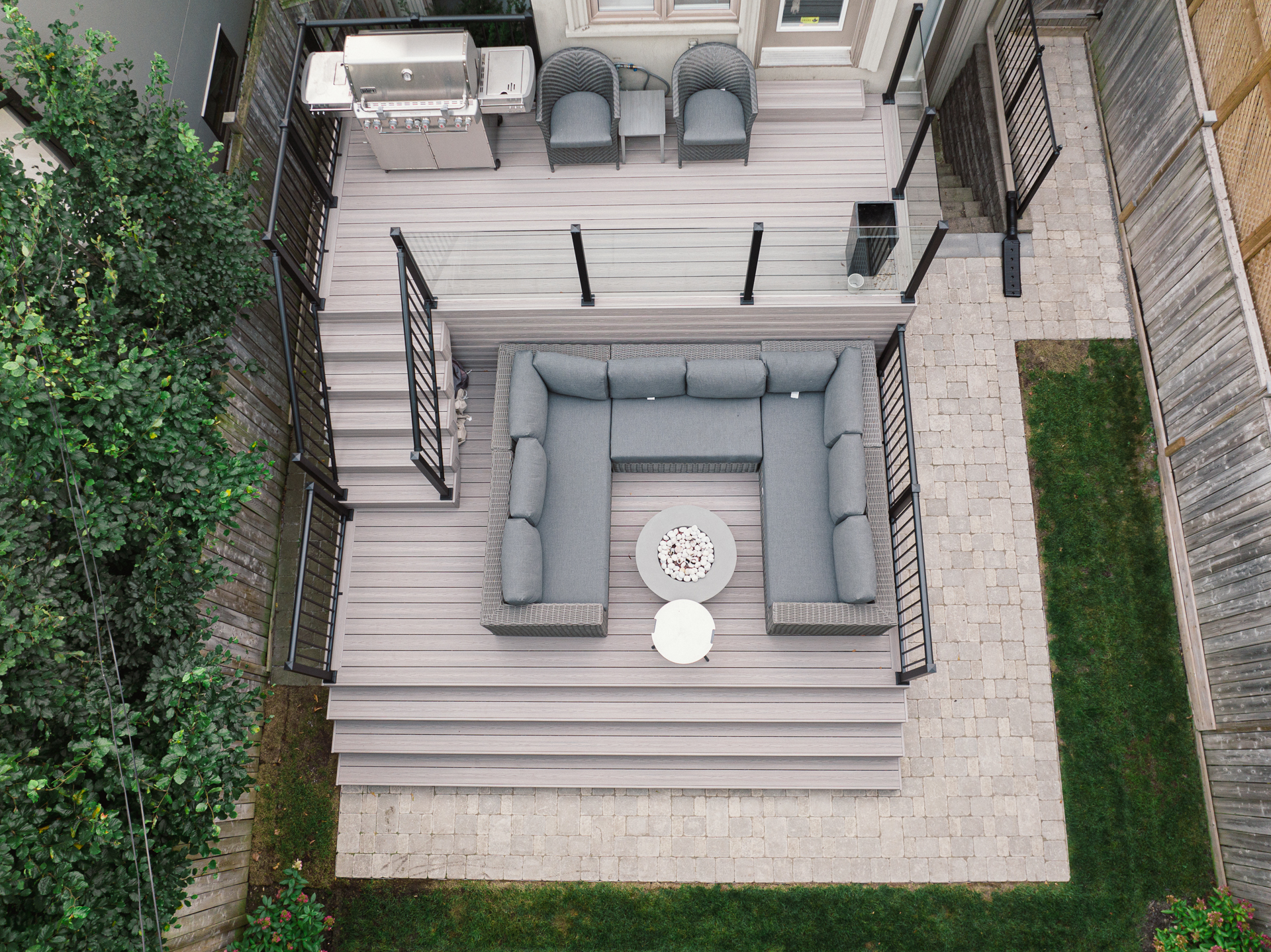 Aerial view of a modern backyard deck with Fiberon Cabana boards, a gray L-shaped sofa, round white table, two armchairs, barbecue grill, and black railings—overlooking a paved patio and lush green grass.