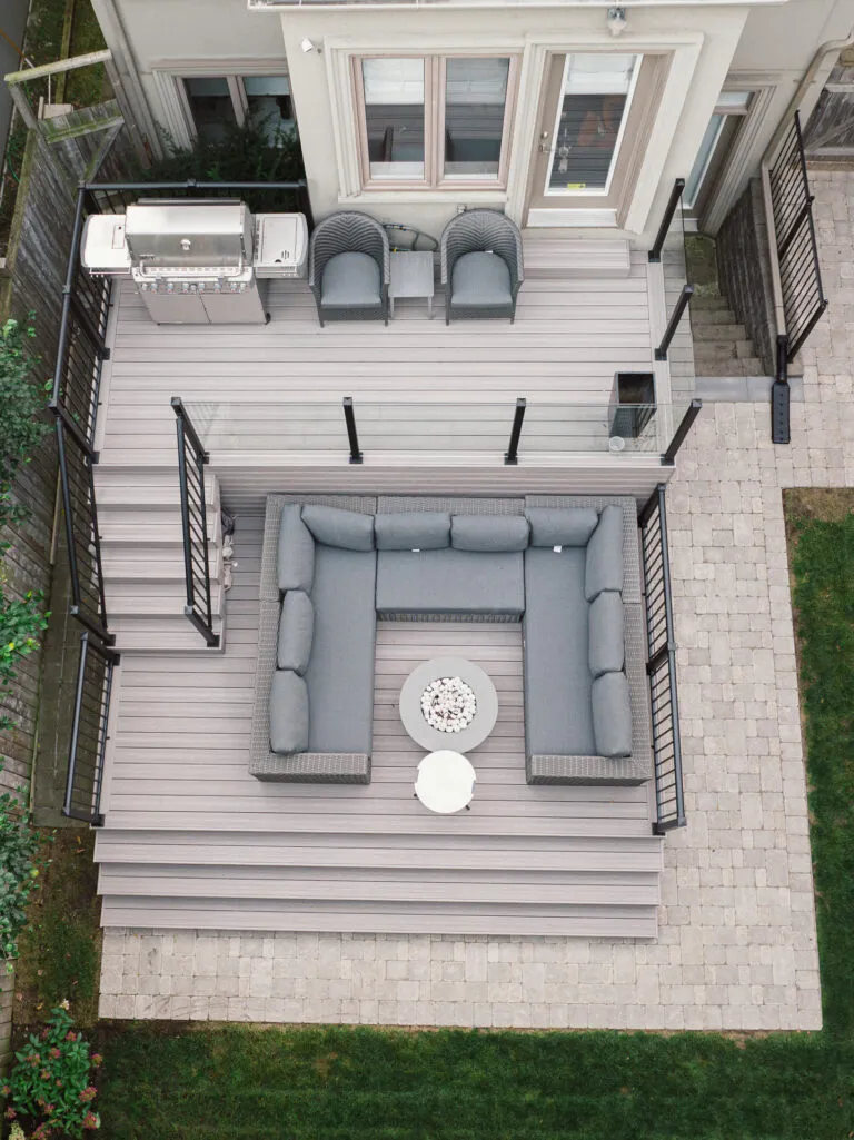Aerial view of a backyard deck with a gray sectional sofa, round fire pit table, two armchairs, and grill. The Fiberon Cabana deck features black railings, stairs down to a stone patio, and a grassy lawn.