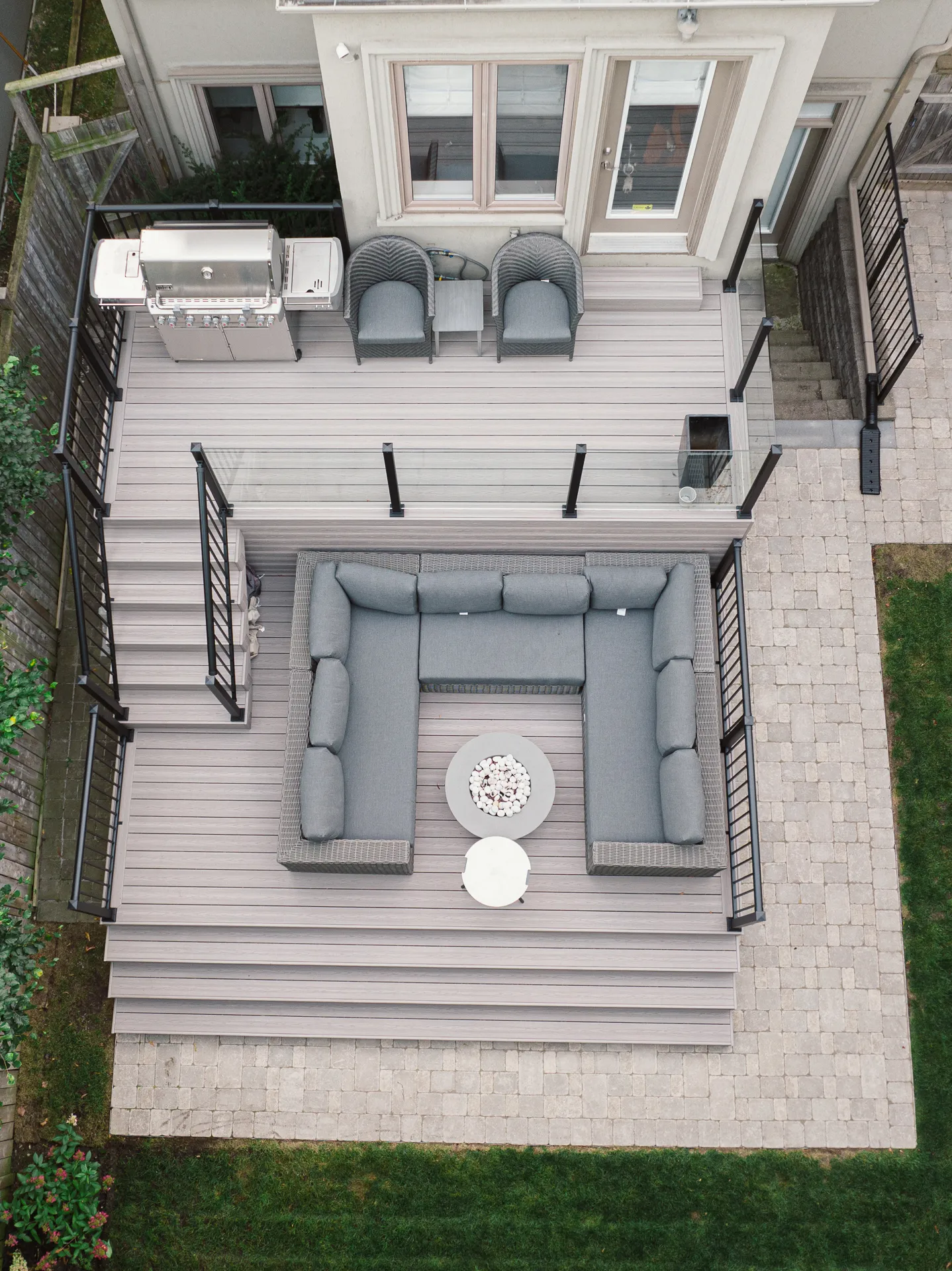 Aerial view of a backyard deck with a gray sectional sofa, round fire pit table, two armchairs, and grill. The Fiberon Cabana deck features black railings, stairs down to a stone patio, and a grassy lawn.