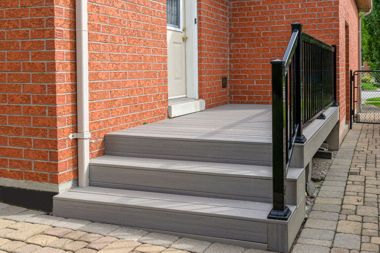 A small gray composite deck with three steps and a black metal railing leads to a white door set in a red brick house. The area is surrounded by interlocking stone pavement.