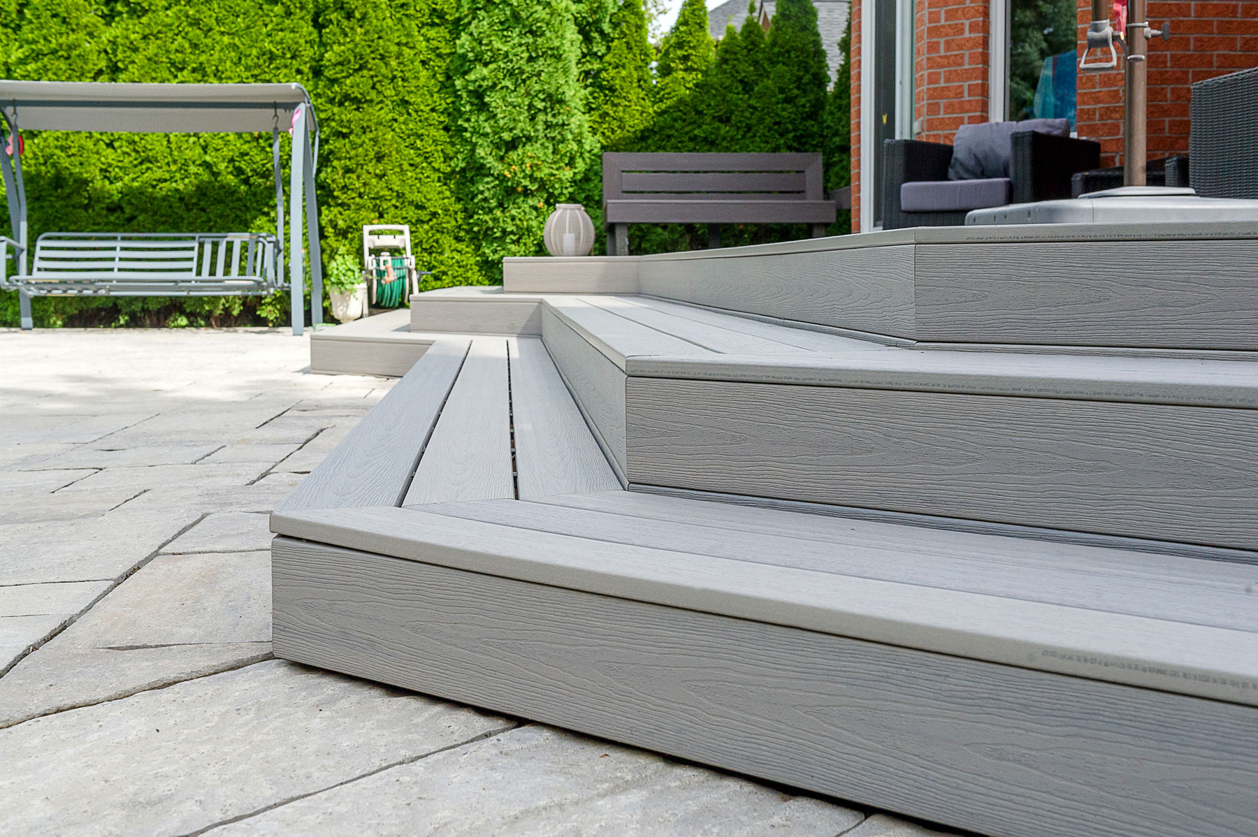 Close-up of wide, light gray composite deck steps leading from a patio to a seating area, with outdoor furniture and green hedges in the background.