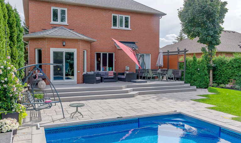 Backyard with a brick house, raised patio with outdoor furniture, red umbrella, and dining area. Steps lead down to a paved area and rectangular swimming pool surrounded by greenery and trees.