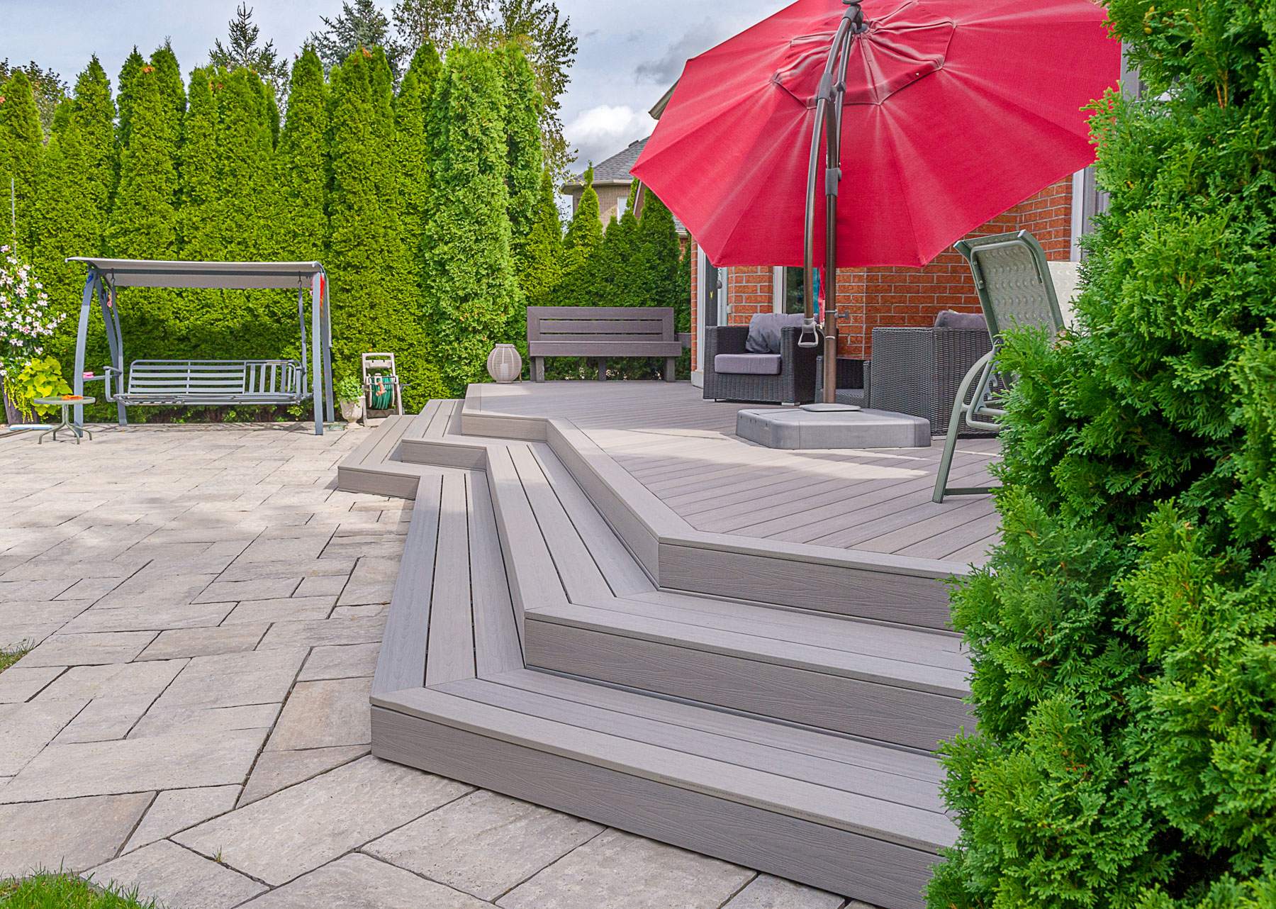 A backyard patio with a raised deck featuring outdoor furniture, a red umbrella, a dining table with chairs, a swing bench, and green shrubs bordering a brick house under a partly cloudy sky.