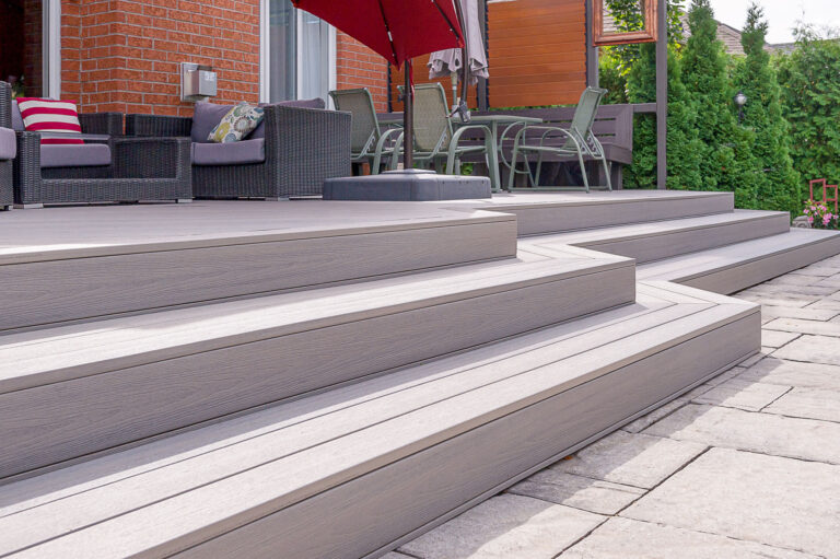 A modern patio with wide, gray composite decking steps leads up to an outdoor seating area with wicker furniture, a glass table with chairs, and a red umbrella, set beside a brick house and green shrubs.