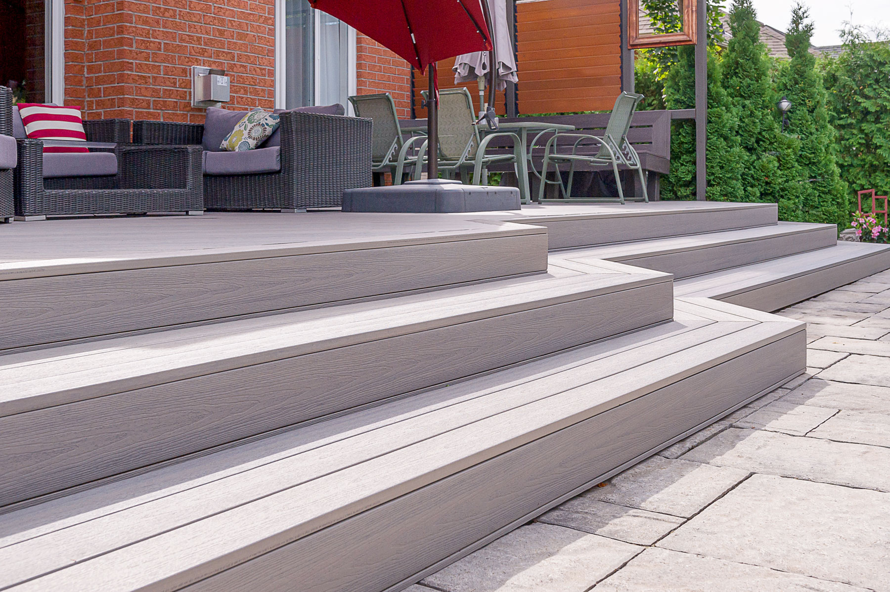 A modern patio with wide, gray composite decking steps leads up to an outdoor seating area with wicker furniture, a glass table with chairs, and a red umbrella, set beside a brick house and green shrubs.