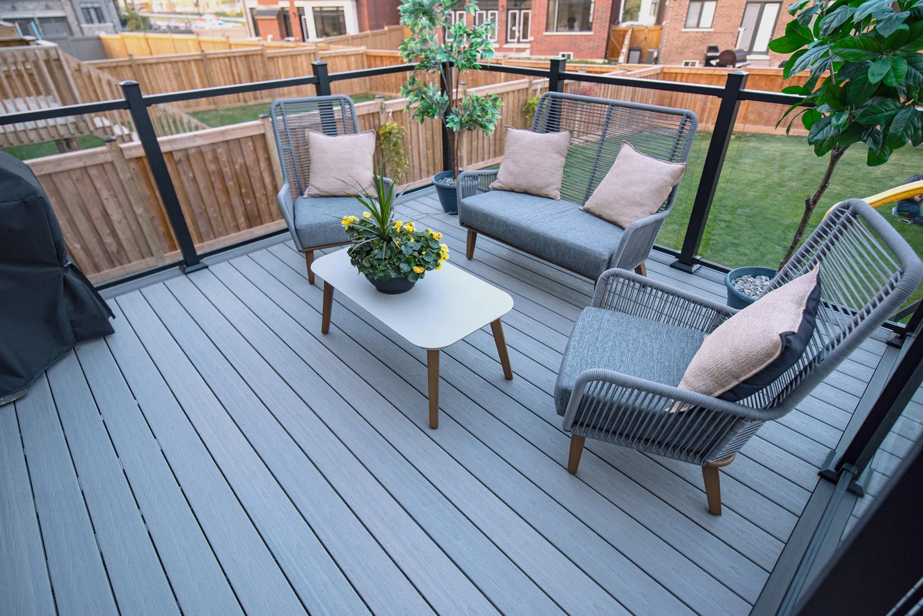 Side-by-side images of the Fiberon modern outdoor staircase with gray steps and black railings leading to a raised patio. The patio is bordered by glass panels and decorated with potted plants. The setting includes brick walls and a paved walkway.