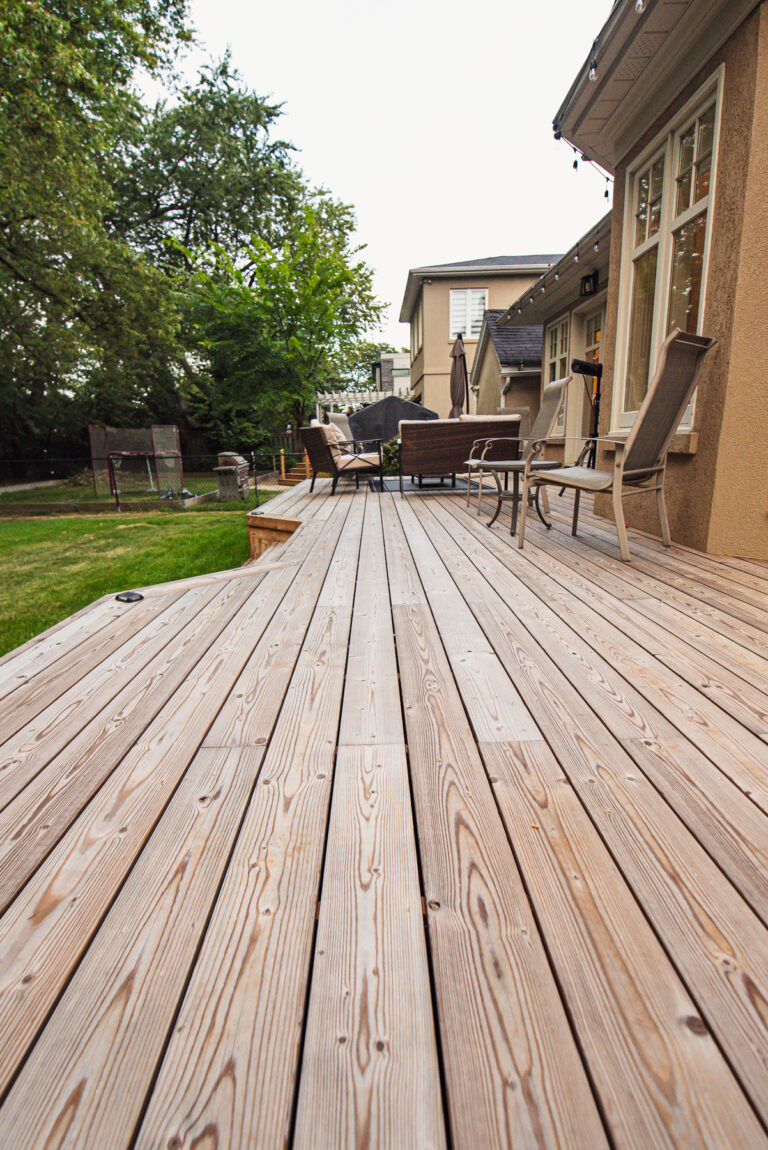 A spacious wooden backyard deck with Lunawood Decking features patio chairs and tables, attached to a house. The deck overlooks a grassy yard with trees and fencing, creating an inviting outdoor seating area.