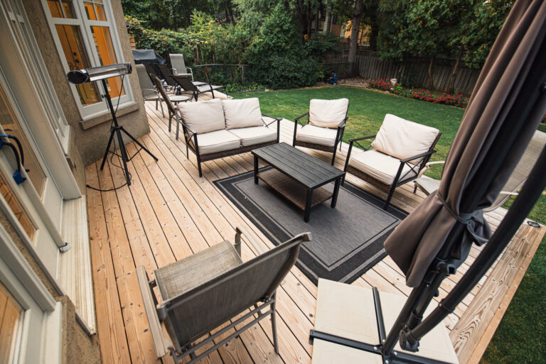 A spacious Lunawood Thermowood Deck with outdoor furniture and a covered grill, attached to a beige house with large windows and a glass door, overlooks a lush green lawn.