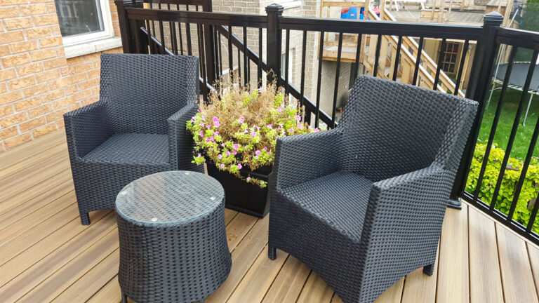 Two dark wicker chairs and a round glass-top table are arranged on a wooden deck next to a large planter filled with blooming flowers, bordered by a black metal railing.