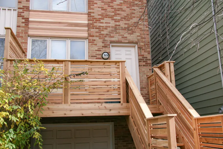 A townhouse with a brick exterior, cedar balcony, and staircase leading to a white front door on the second level.