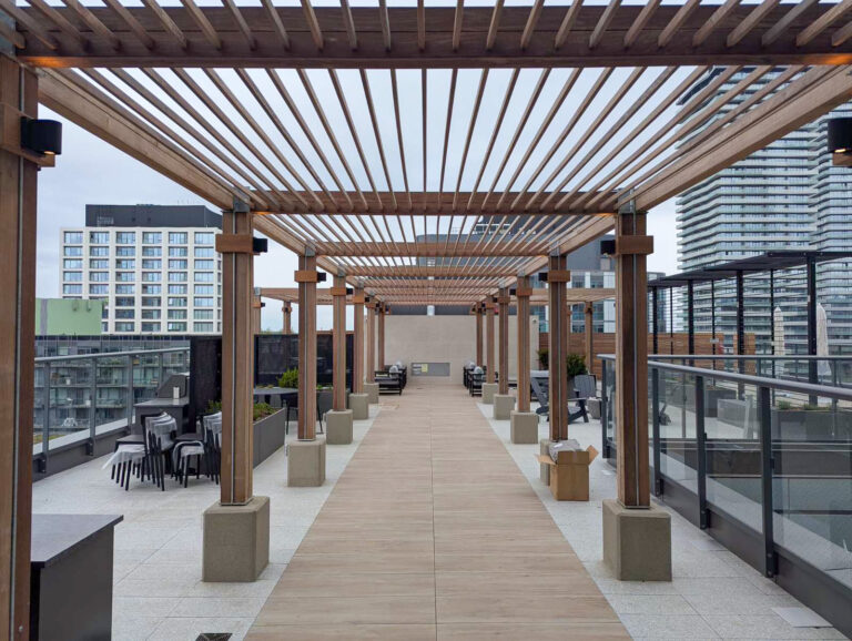 A rooftop patio with wooden pergolas, seating areas, and city buildings visible in the background under a cloudy sky.