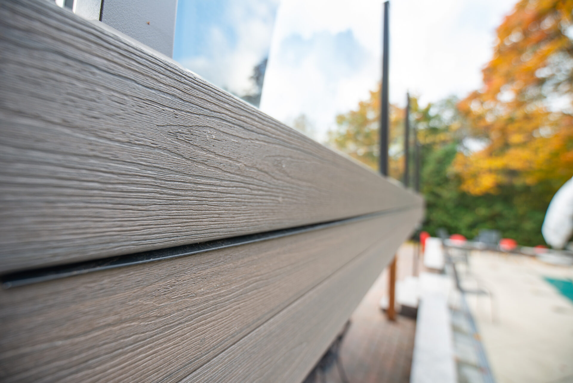 Close-up of horizontal wooden panels, resembling Fiberon composite decking, on an outdoor wall with a blurred background featuring autumn foliage and a swimming pool area.