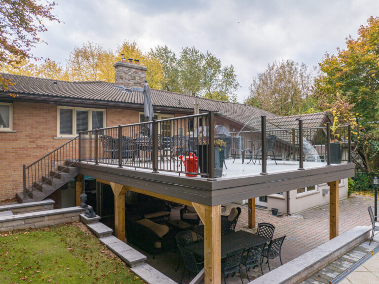 Two-level outdoor deck attached to a brick house. The upper deck has glass railings and patio furniture, while the lower area features a covered seating space with a dining table and chairs. Trees with autumn leaves surround the backyard.
