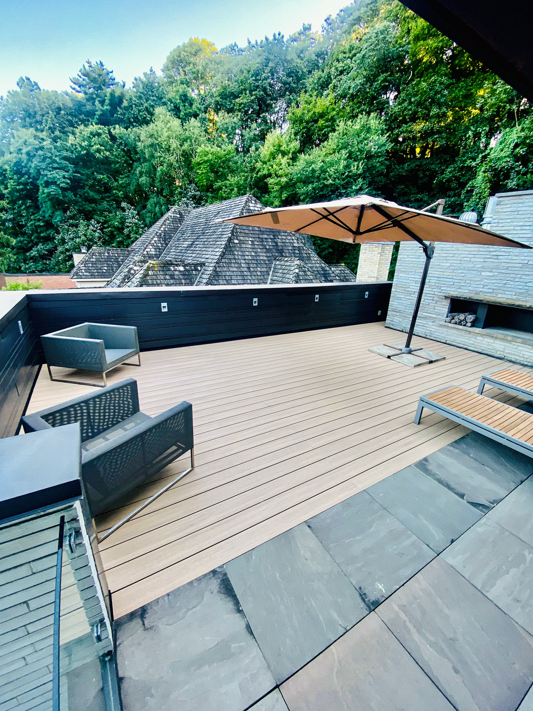 A modern rooftop patio features Azek PVC decking, outdoor seating, a large umbrella, and a forested backdrop.