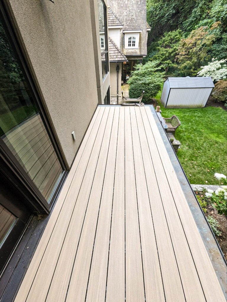 Light-colored wooden deck outside a house, overlooking a backyard with trees, a shed, and a neighboring house.