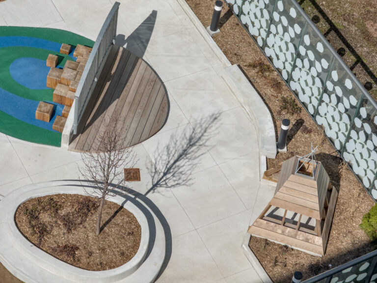 Aerial view of a playground with wooden structures, blue and green rubber flooring, and landscaped areas.