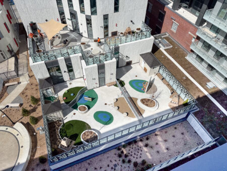 A rooftop playground with artificial grass, seating areas, slides, and shade structures, viewed from above.