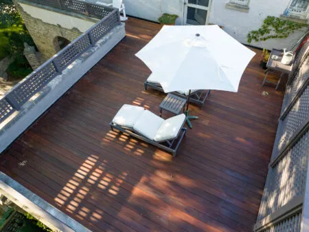 Aerial view of an IPE deck featuring a white patio umbrella and reclining chairs, complemented by additional seating along the perimeter. The area is framed by a gray railing, with lush green trees providing a serene backdrop.