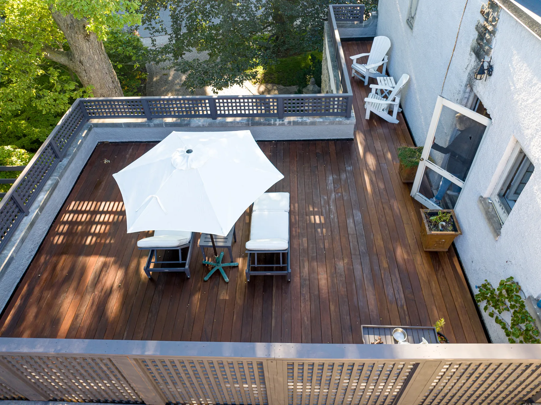 Aerial view of an IPE deck featuring a white patio umbrella and reclining chairs, complemented by additional seating along the perimeter. The area is framed by a gray railing, with lush green trees providing a serene backdrop.