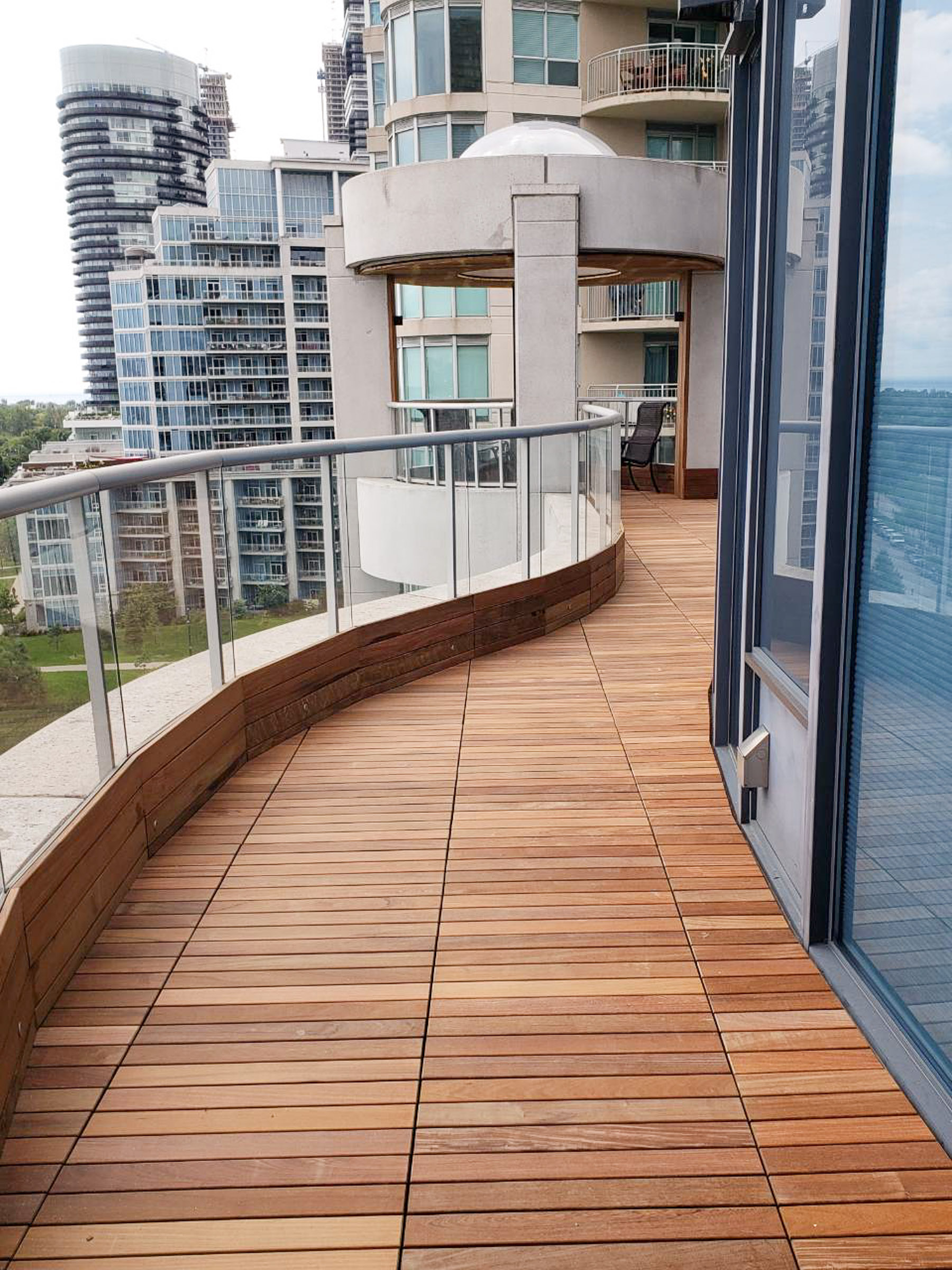 Curved IPE wood balcony with glass railing on a high-rise, overlooking modern city buildings in the background.