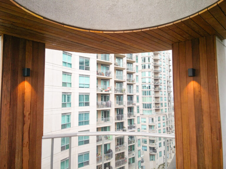 View of a modern apartment building through a balcony framed by wood paneling and wall lights.