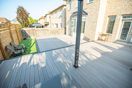 A spacious backyard deck with light wood flooring, fenced yard, two chairs on artificial grass, and part of a brick house.
