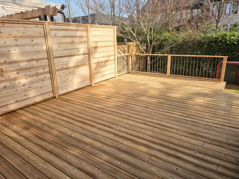 A spacious wooden deck with privacy screens on the left and a railing on the right, overlooking a backyard with trees and shrubs, in a residential area.