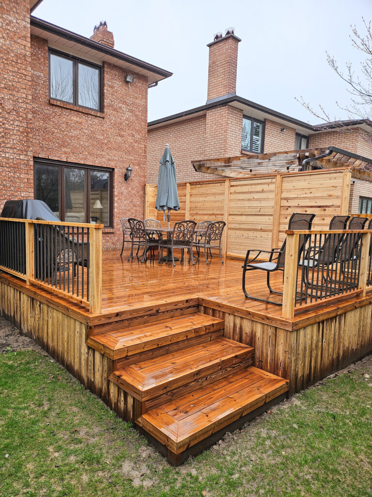 A freshly stained Lunawood Decking backyard deck with steps, outdoor dining table, chairs, grill, and umbrella, attached to a brick house. A privacy screen separates the deck from the neighboring yard.