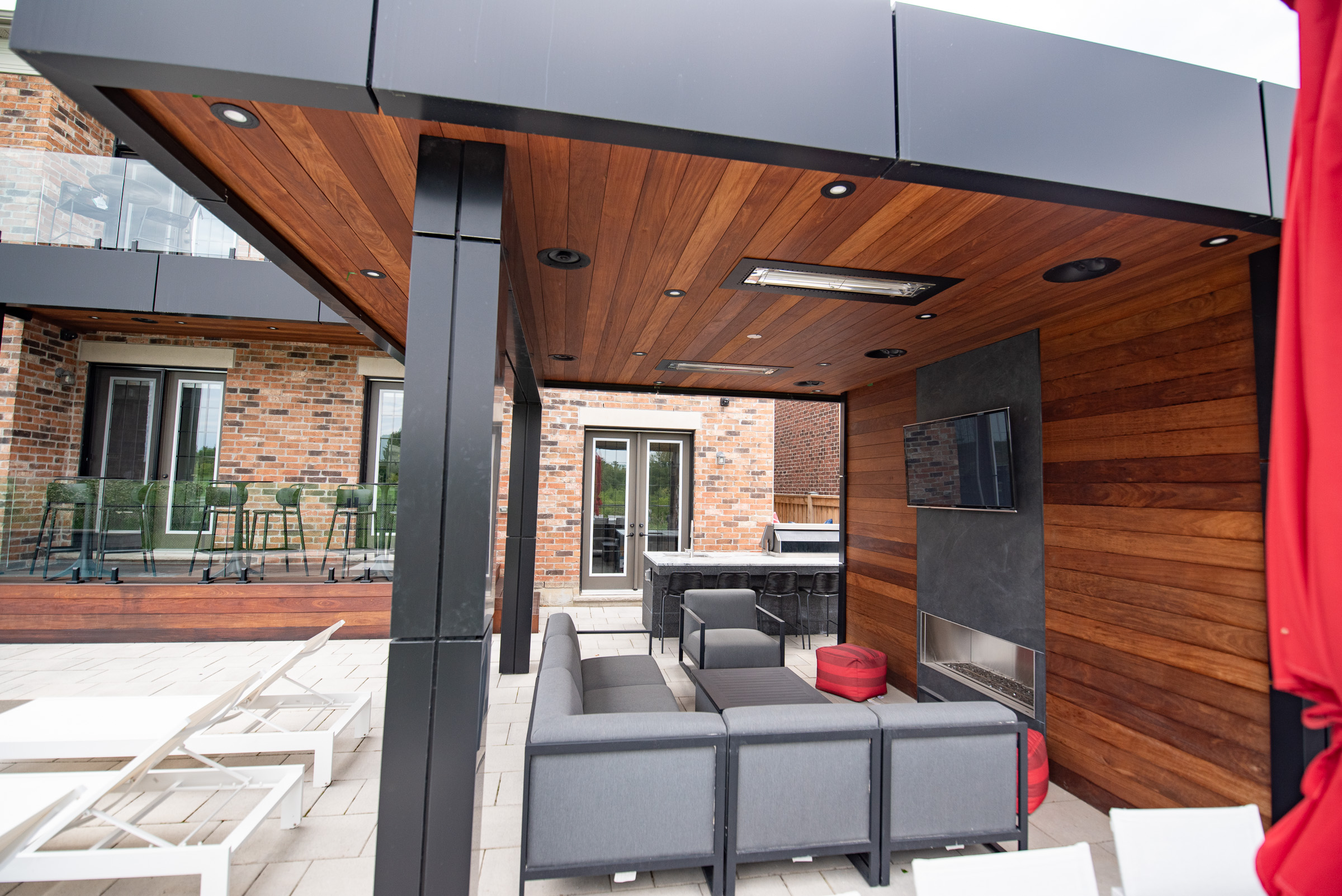 Covered outdoor patio with wood ceiling and walls, lounge seating, TV, fireplace, and a bar area, next to a brick house.