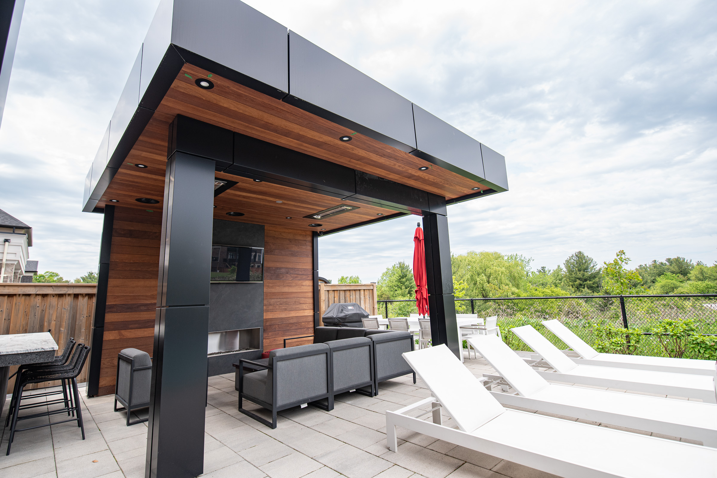 Modern outdoor patio with a wood-paneled pergola, gray seating, lounge chairs, and a red umbrella on a paved surface.