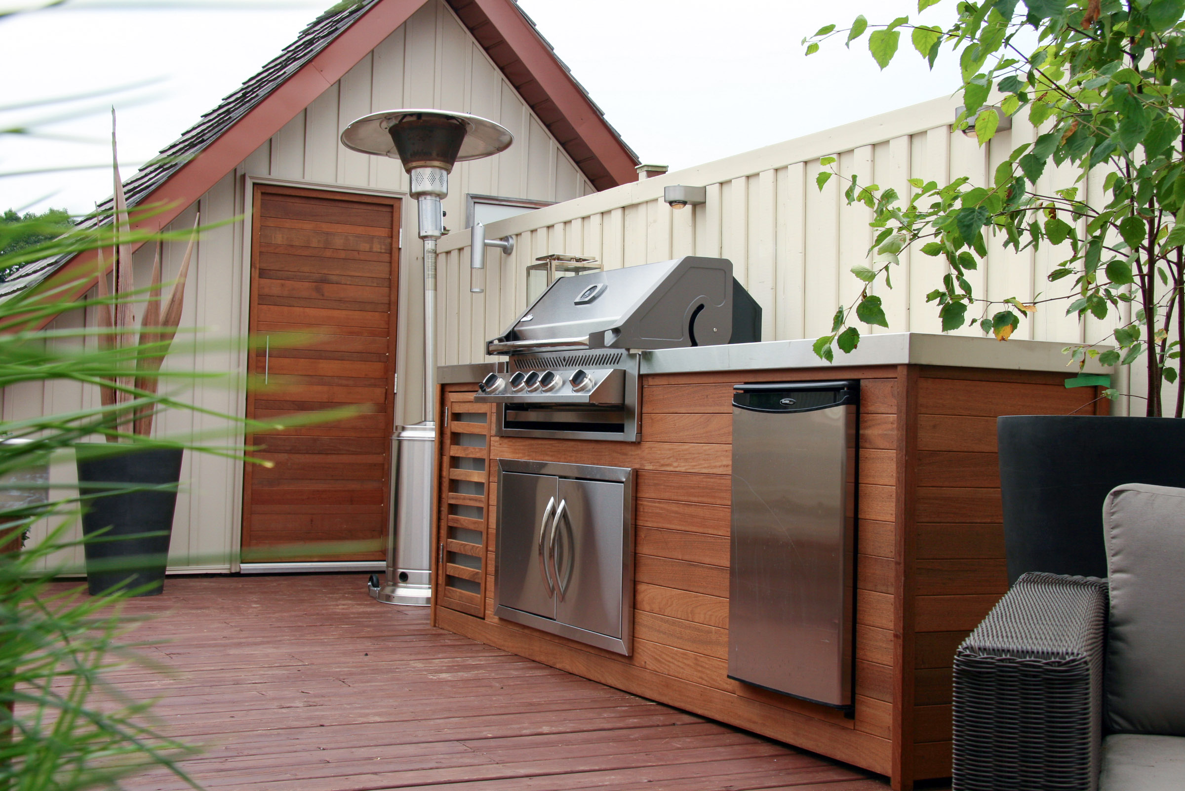 Outdoor patio with a stainless steel grill, mini fridge, heater, and wooden cabinetry next to a small house.