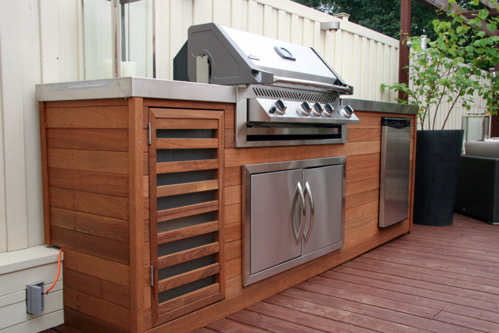Outdoor kitchen setup with a built-in stainless steel grill, storage cabinets, and a mini fridge on a wooden deck.