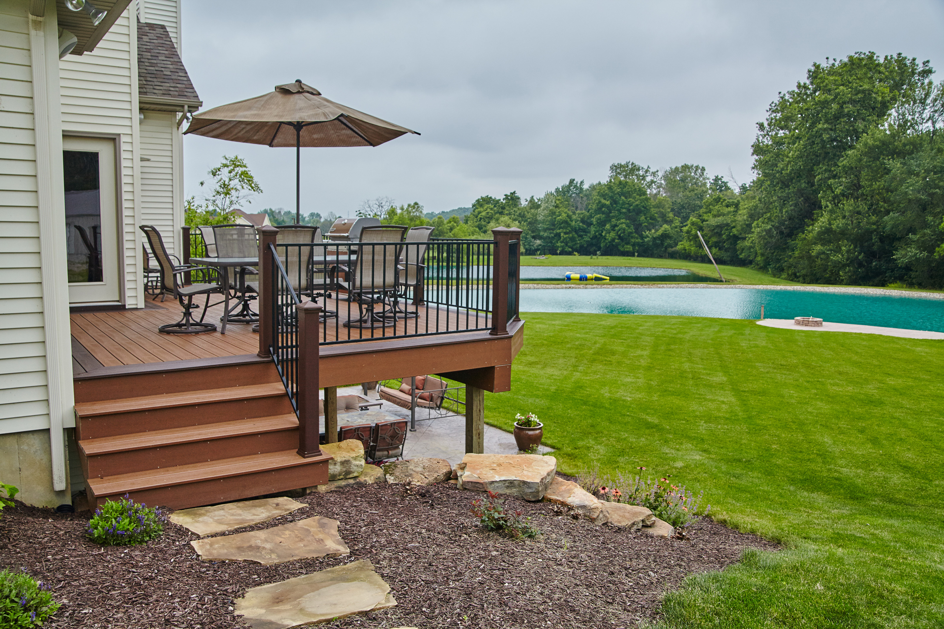 A wooden deck with outdoor dining furniture and an umbrella overlooks a green lawn and a pond, surrounded by trees. Stone steps lead from the deck to the yard, and a patio is partially visible below.