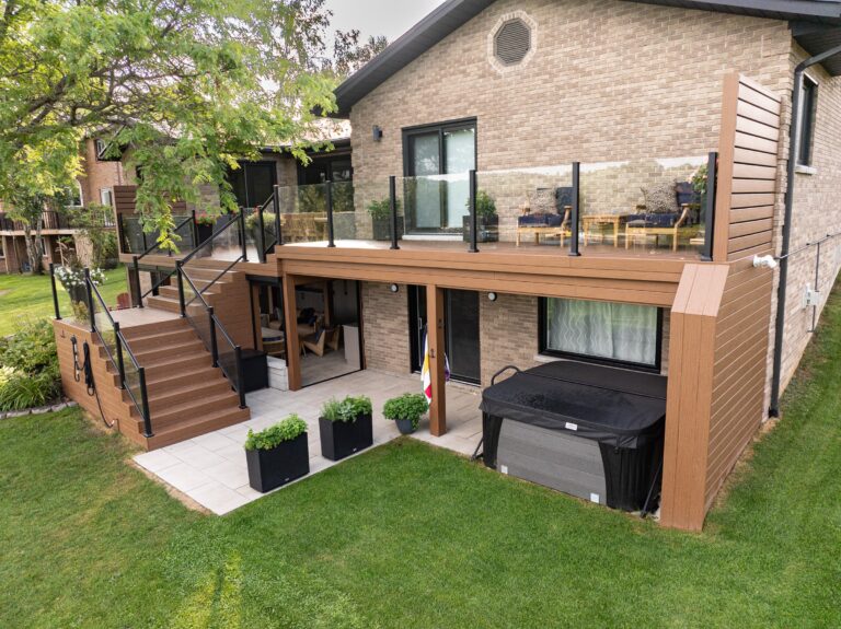 Two-level deck with glass railing, outdoor seating, covered patio, hot tub, and landscaped yard next to a brick house.
