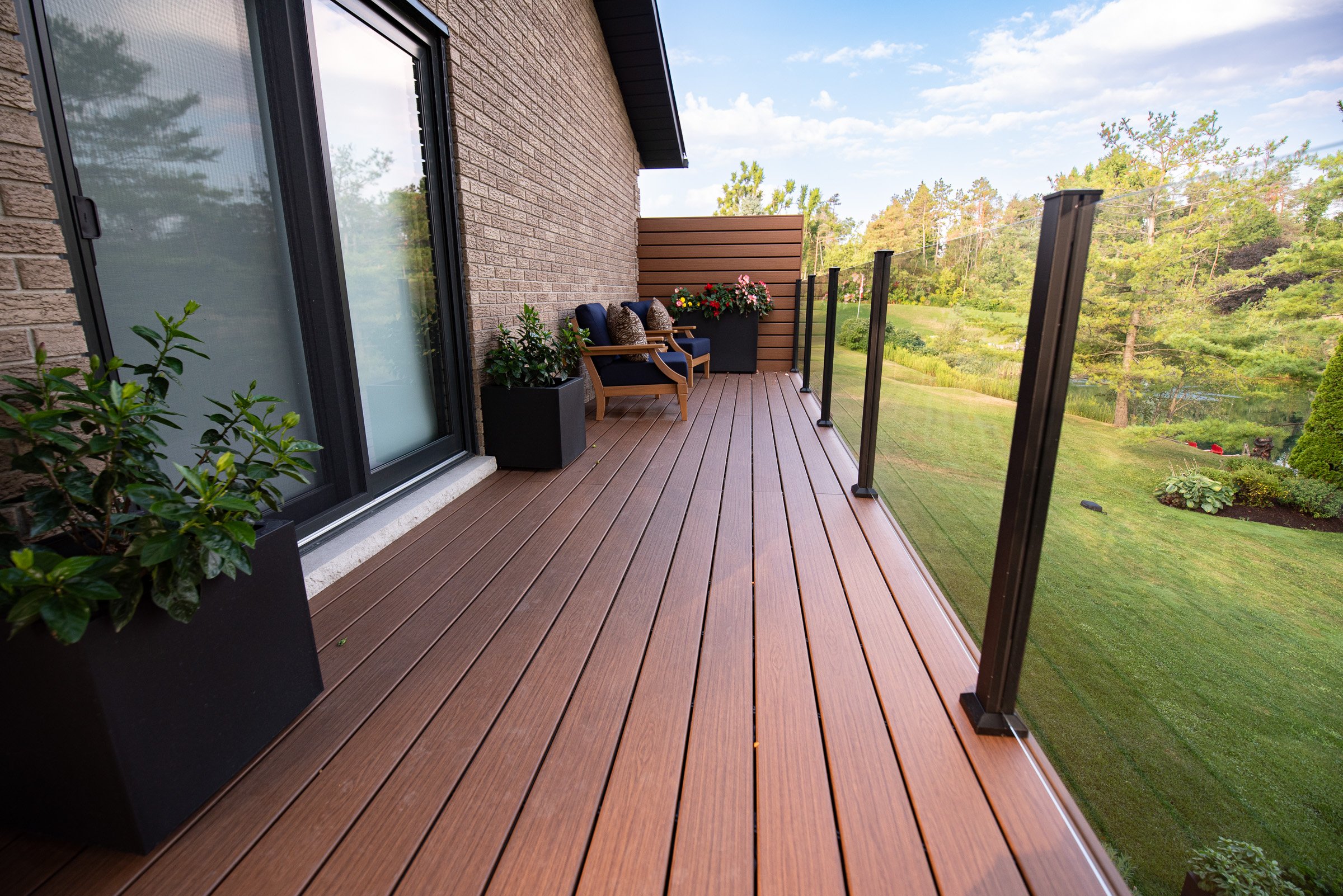 A modern deck with glass railing, potted plants, and outdoor seating overlooks a green lawn and trees.