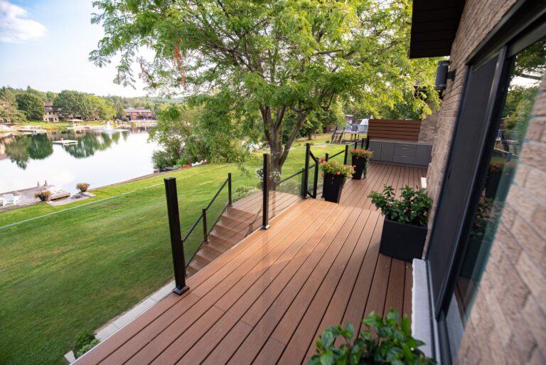 A wooden deck with glass railing overlooks a grassy yard and a calm lake, surrounded by trees and houses.